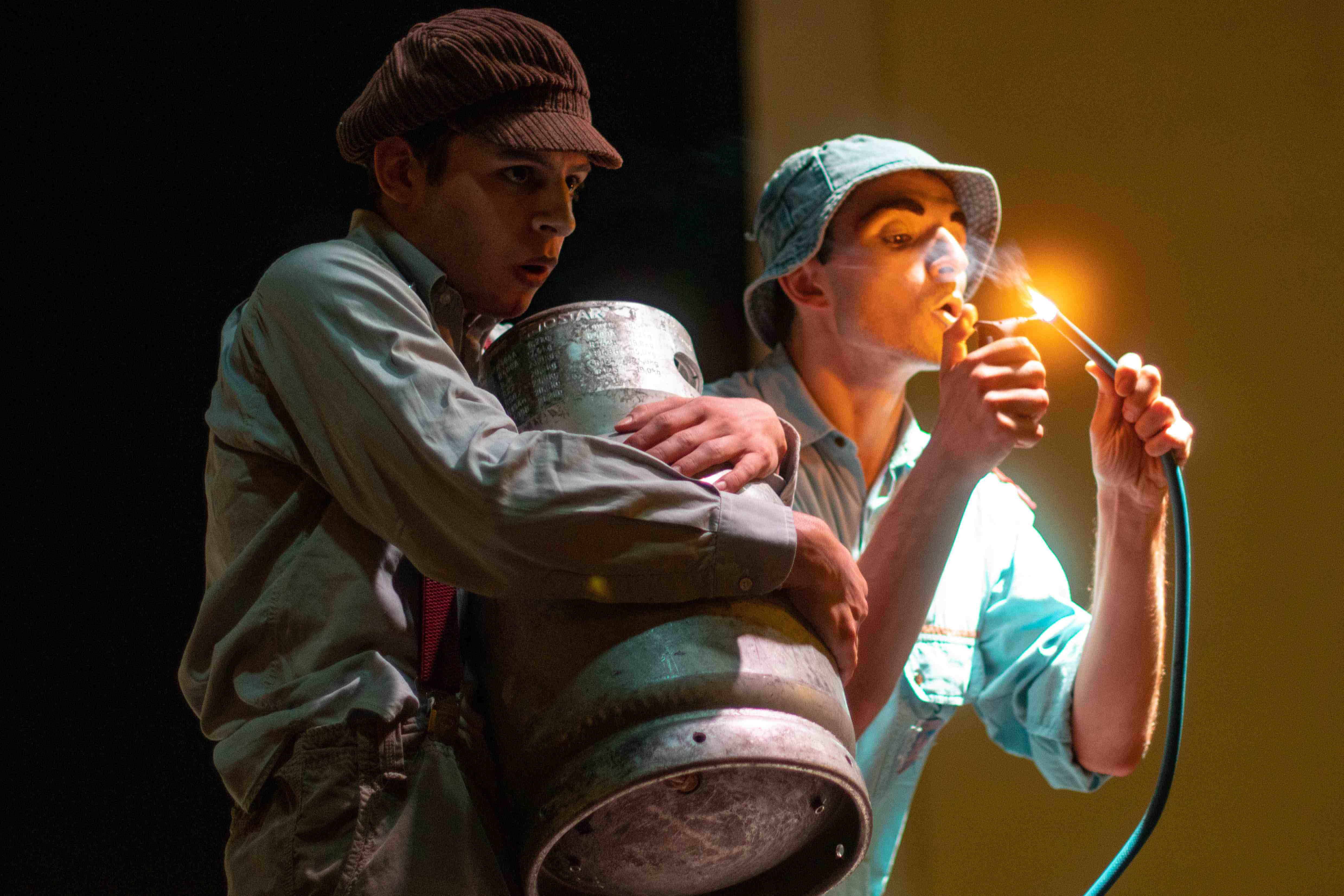 A clown holds a gas tank while another one lights a fire from a plastic tube.