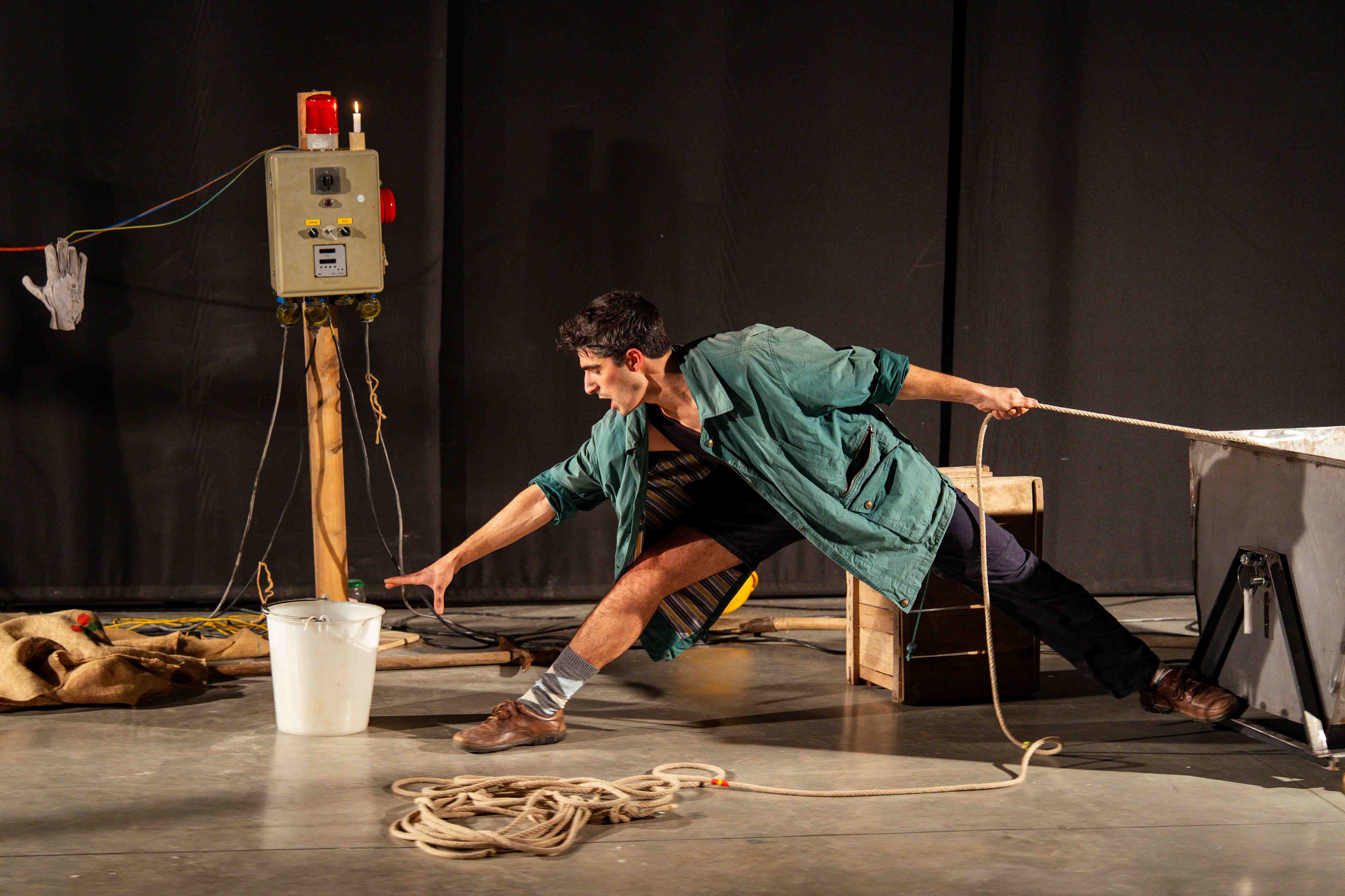 Un clown in giacca verde accovacciato tira una corda, circondato da attrezzi da miniera e un secchio.