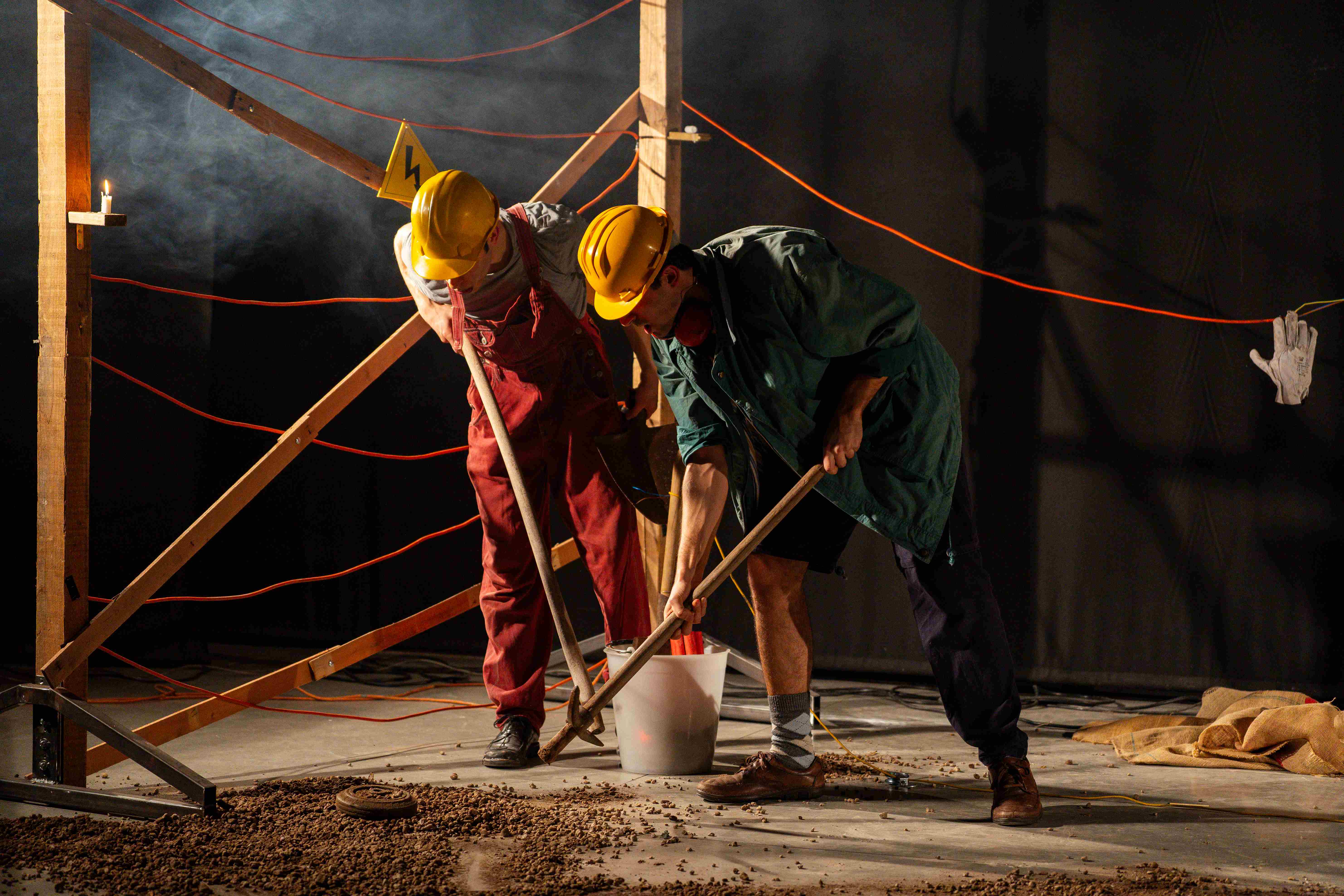 Due clown con caschetti gialli scavano con le pale in una miniera, circondati da impalcature e cavi rossi.