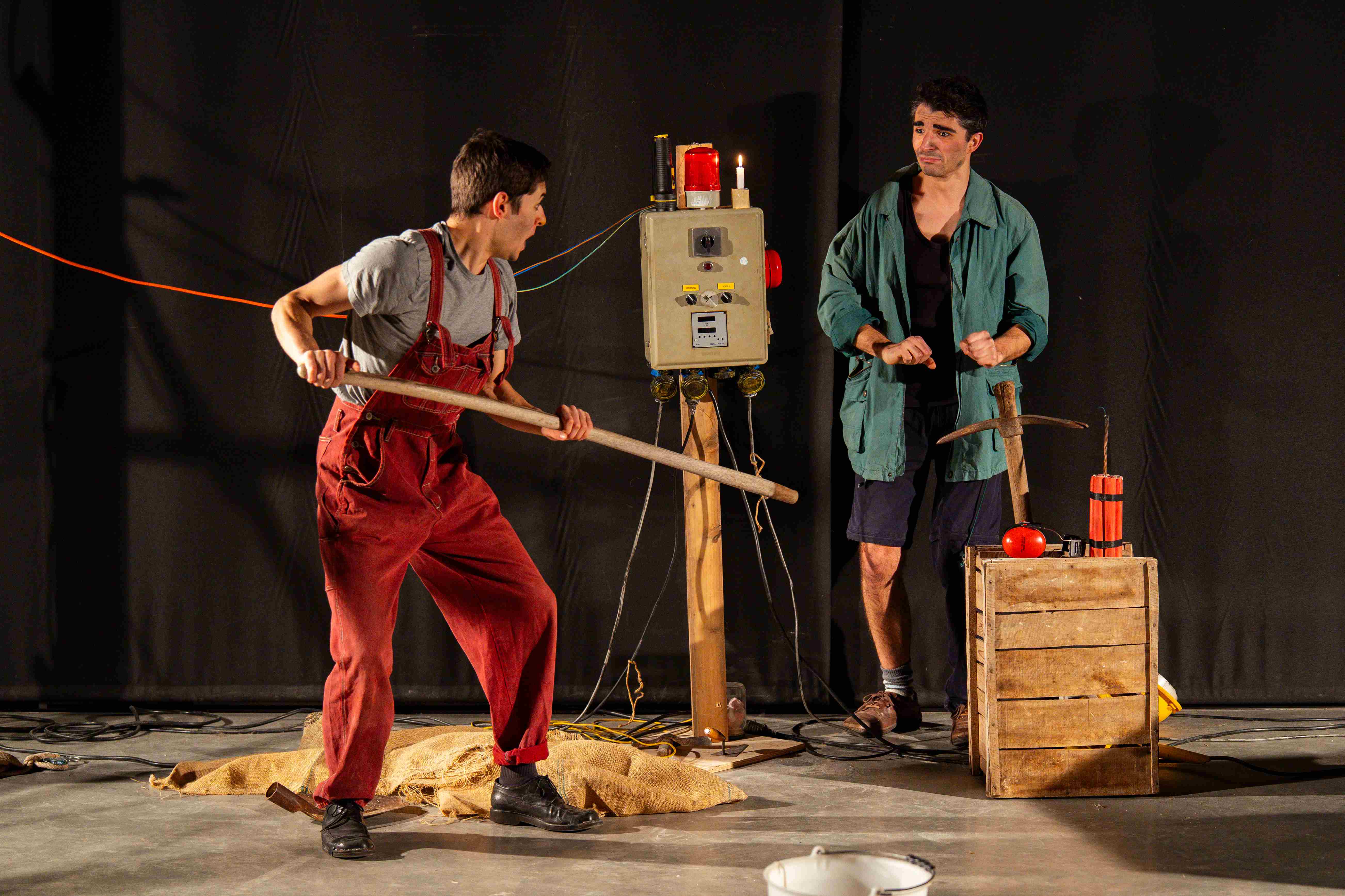 Un clown in tuta rossa impugna un bastone mentre l'altro in giacca verde osserva, tra un quadro elettrico e una cassa di legno.