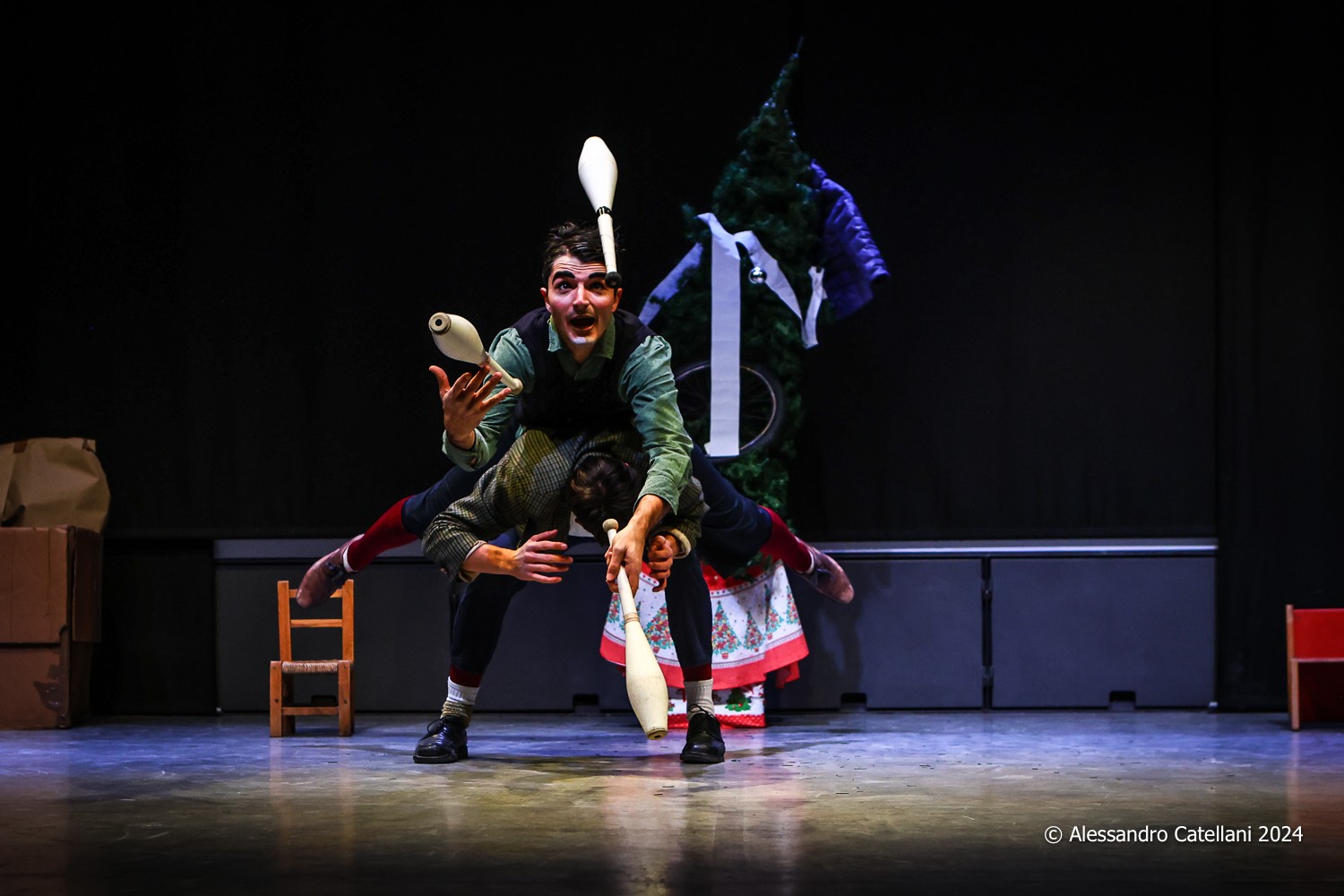 A clown juggles with three clubs lying on another clown's back.