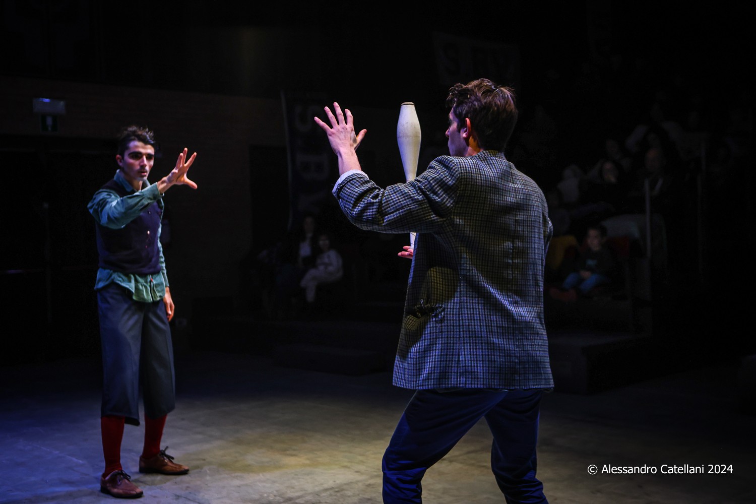 One clown waves his hand in front of another clown with a club, with the audience in the background.