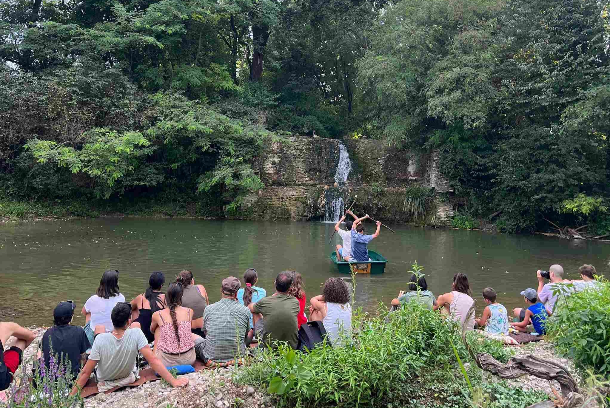 Audience sitting on the shore watches the two actors paddle in the middle of the pond.