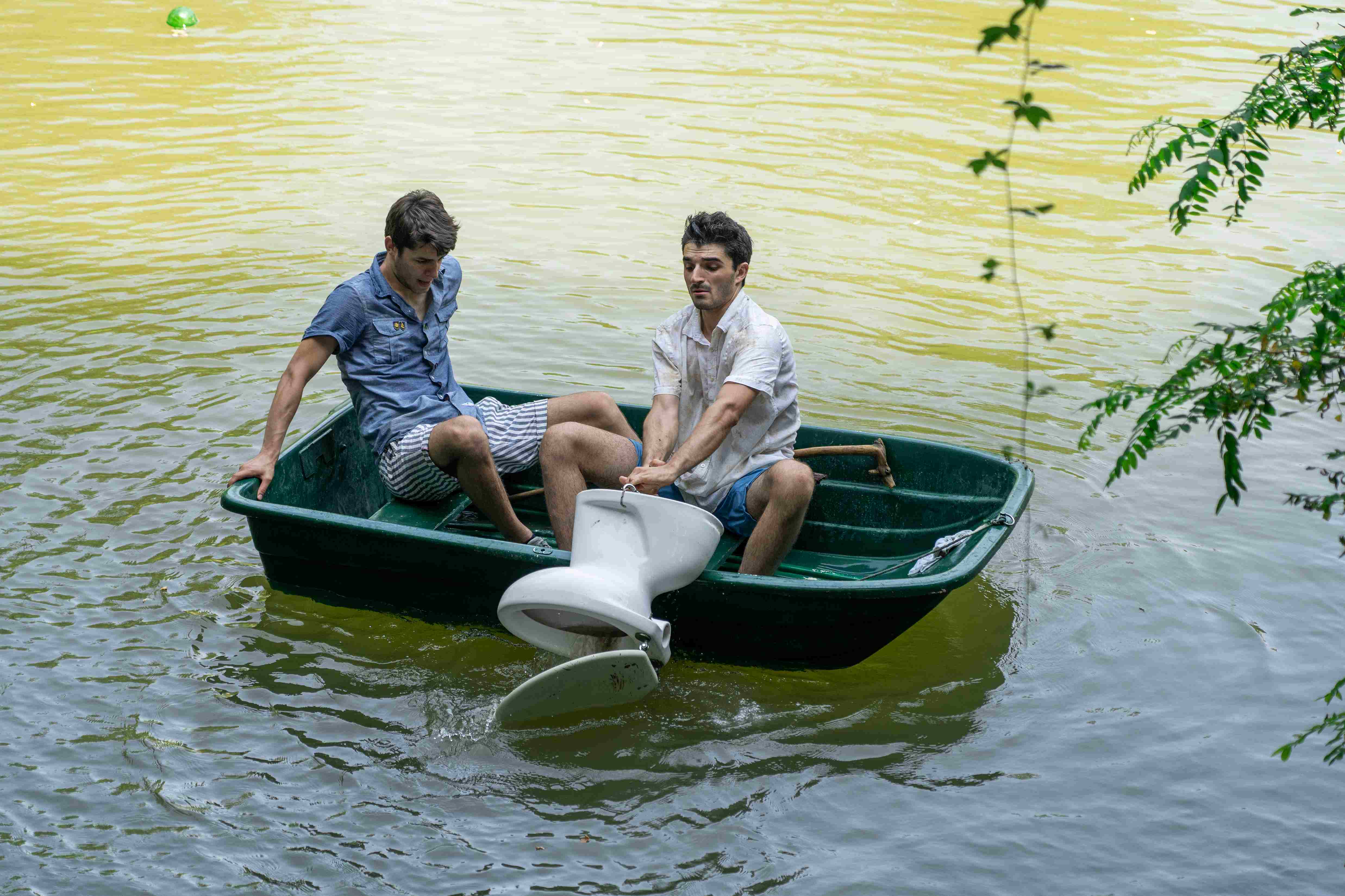 Two clowns in a small boat retrieve a toilet from the bottom of the lake.