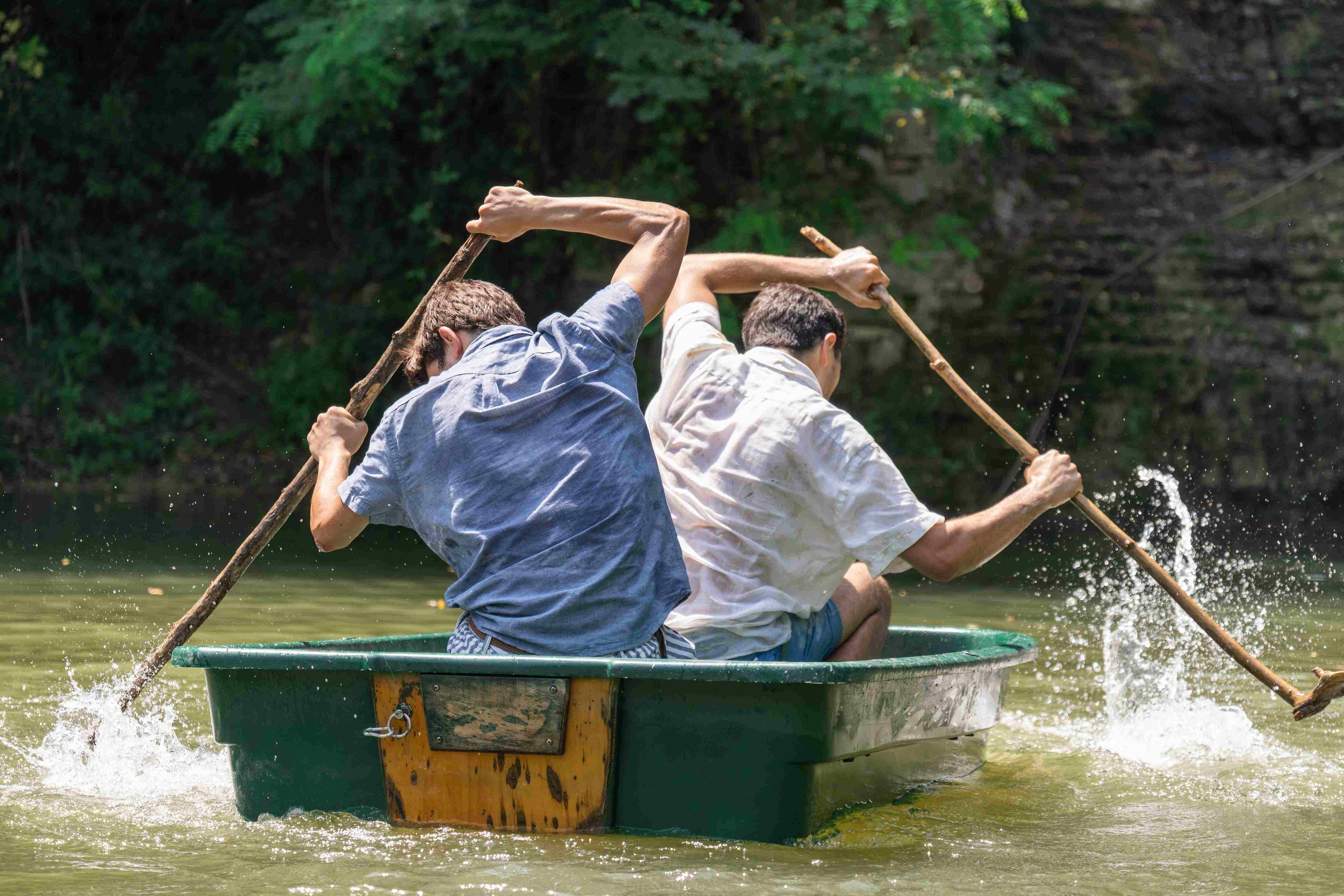 Two characters in a little green boat row with sticks.