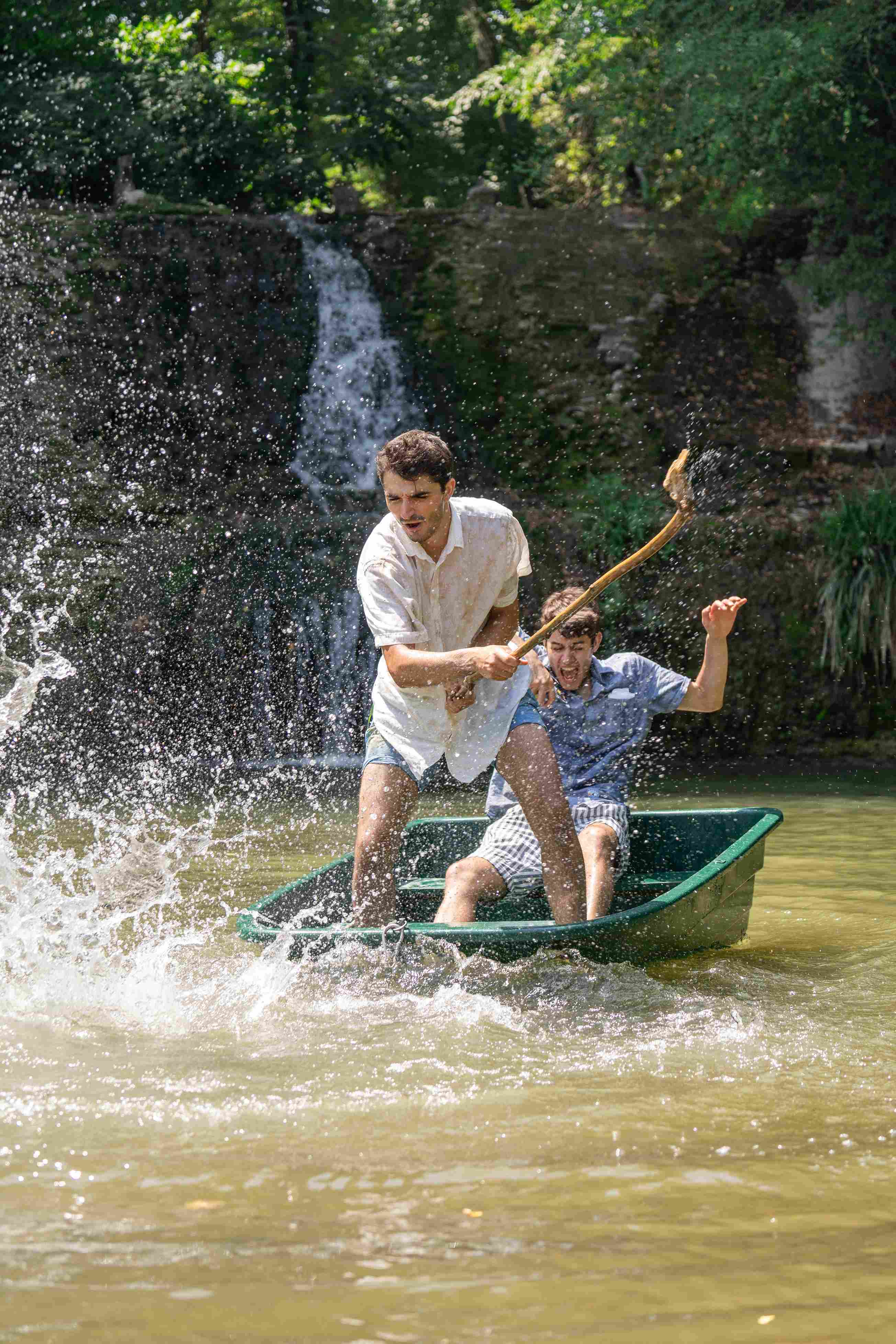 A clown standing on a small boat splashes the water with a stick while another is sitting behind amused.